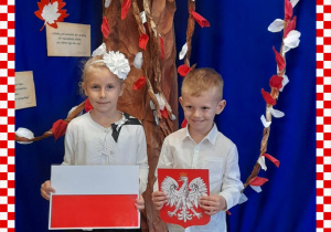 Hania i Kacper w odświętnym ubraniu trzymają w rączkach godło i flagę Polski, w tle dekoracja okolicznościowa
